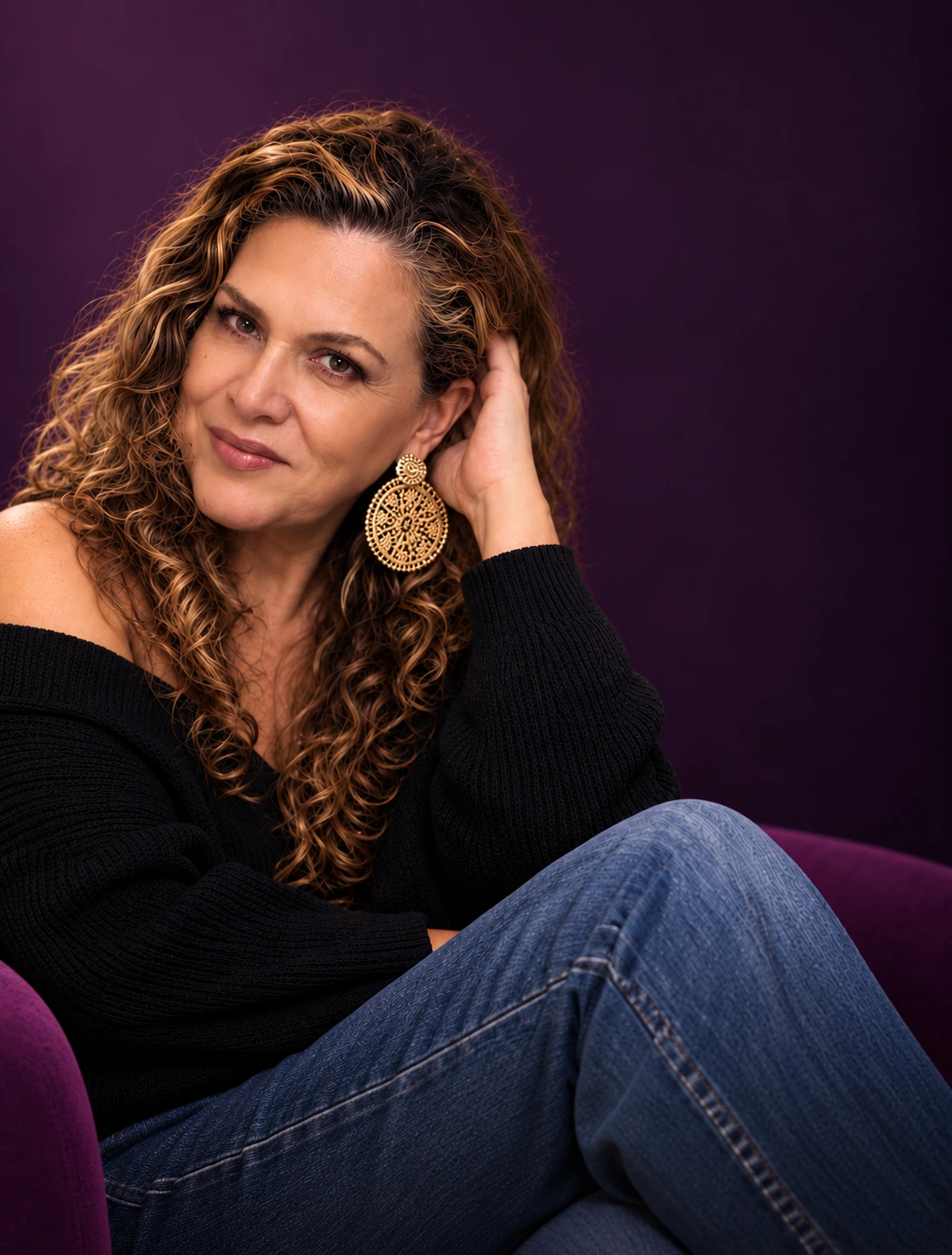 Sandra Echeverri sonriendo, sentada en un sillón morado, con suéter negro y aretes dorados